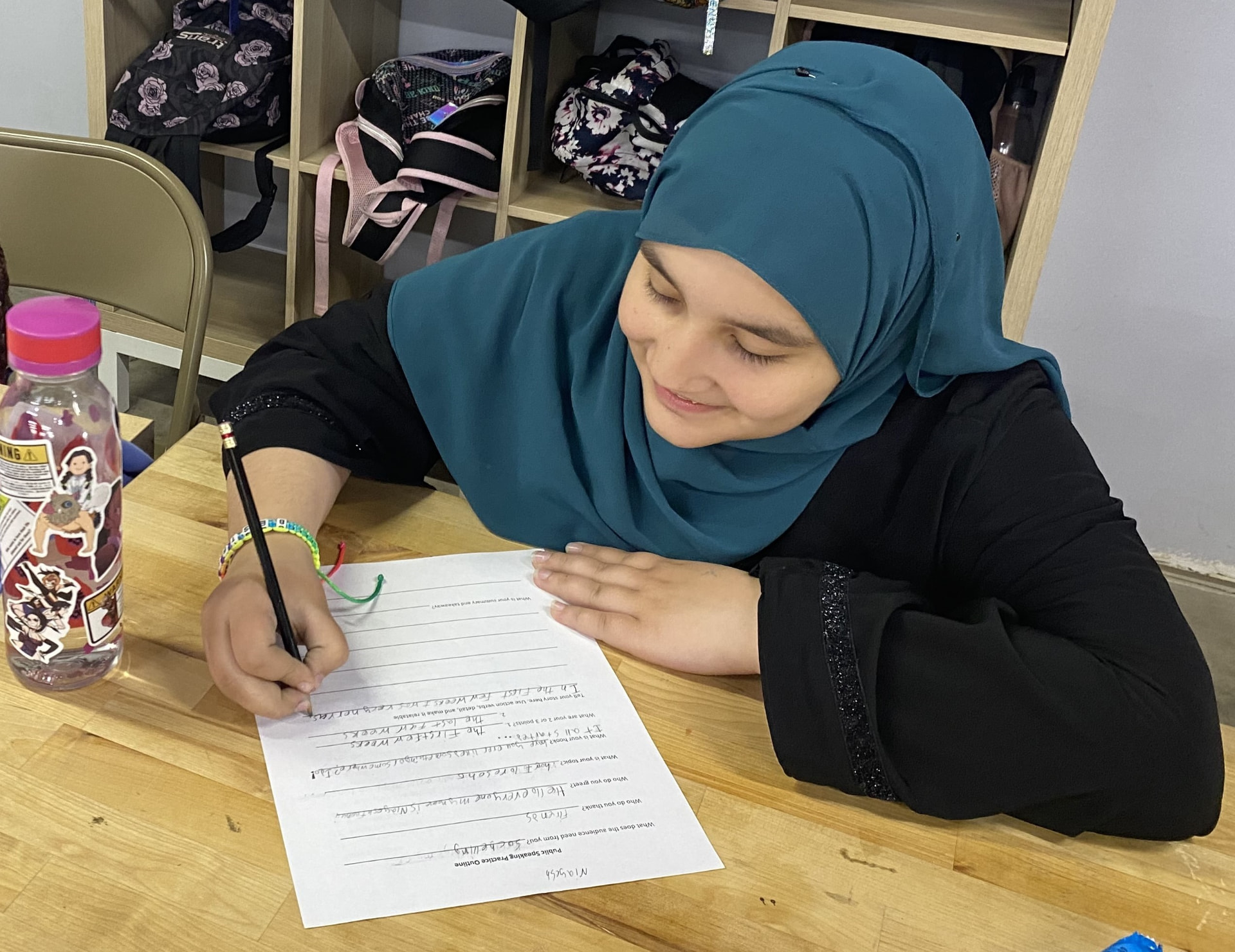 A teenage participant wearing a teal hijab writes on a public speaking practice outline at a workshop table.